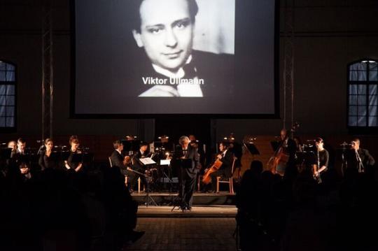 conductor standing in front of ensemble in front of slide show with picture of Viktor Ulmann